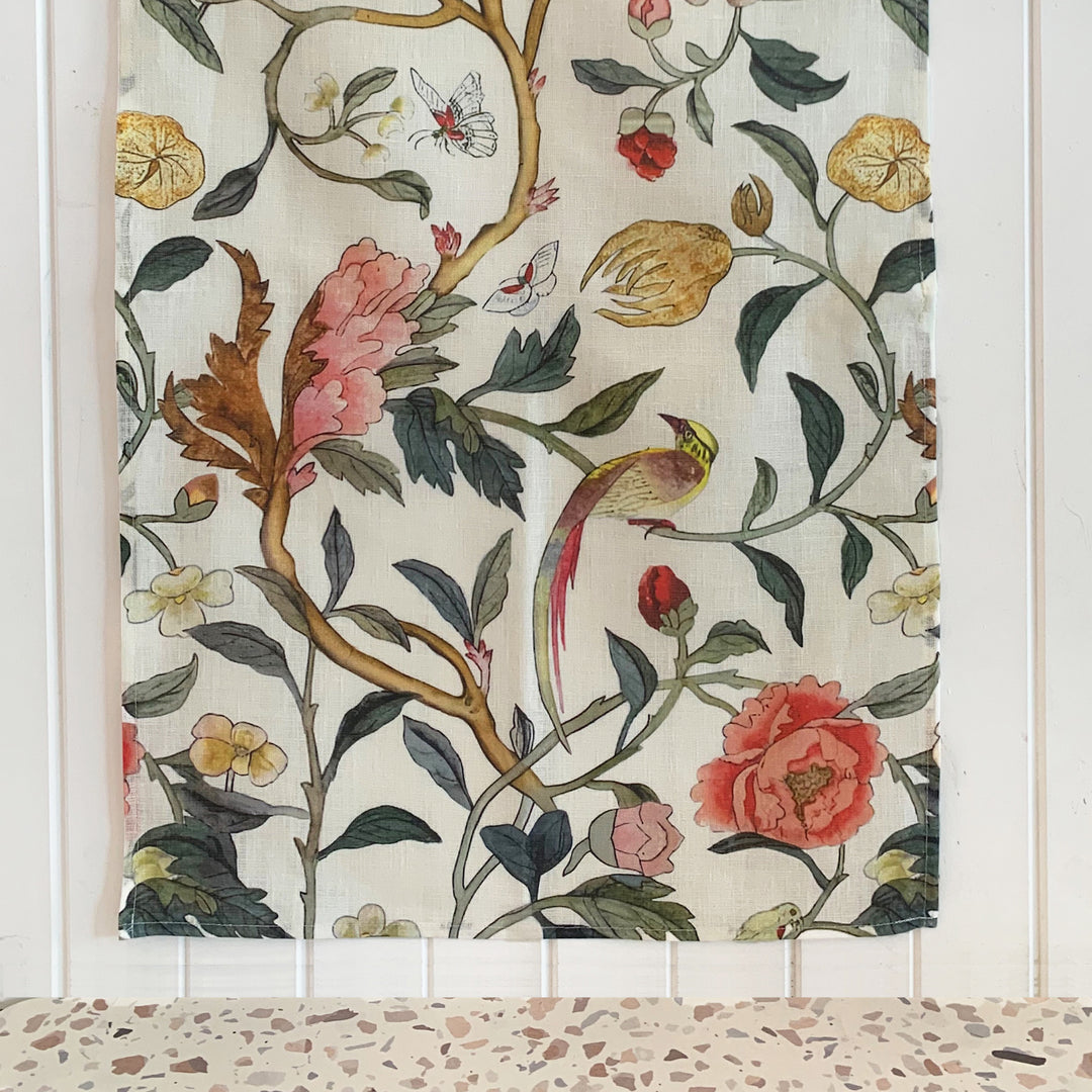 A linen tea towel with a vintage floral pattern including soft pink peony blooms, playful birds, and butterflies, displayed on a white surface.