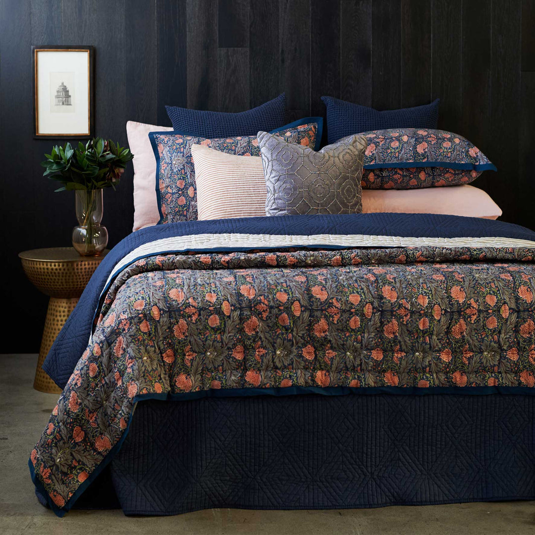 A bed with a floral coverlet set in shades of gray and pink, including two standard shams. The coverlet is displayed on the bed with a dark headboard in the background and a bedside table with a plant and lamp to the side.