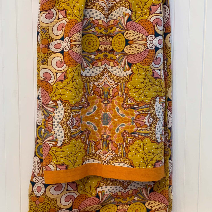 A folded view of the Daphne blanket showcasing the vibrant ochre side with mustard leaves, coral accents, and concentric circle motifs. Hangs neatly from a white wall rail.