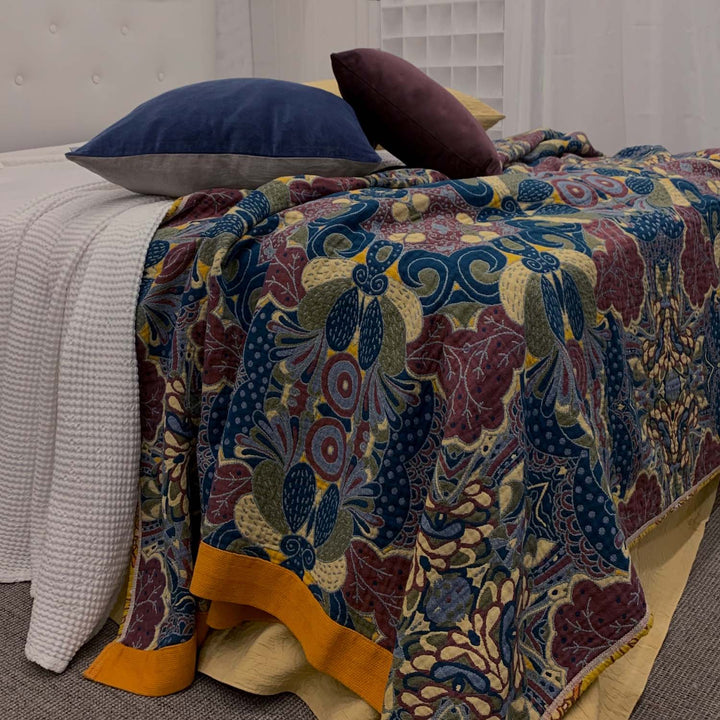 The Daphne blanket shown with the reverse side facing up, featuring cool navy, olive, and burgundy tones. Styled with white bedding and velvet cushions in navy and plum for a moody, layered look.