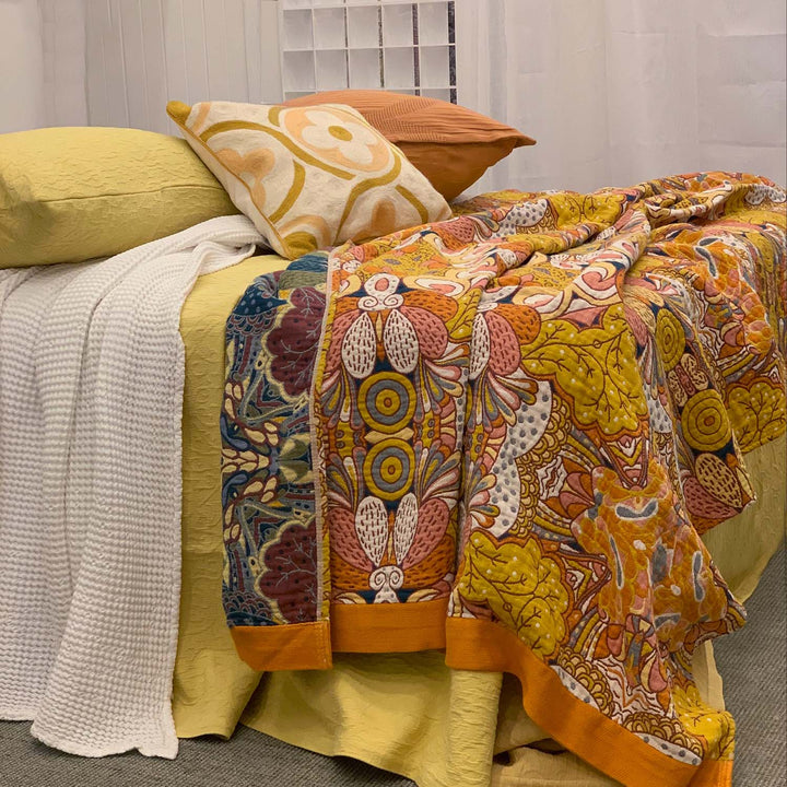 A close-up of the Daphne woven cotton blanket in warm ochre, rust, and mustard tones styled on a yellow bed with a white waffle throw and retro-inspired cushions. The bold floral and abstract pattern adds rich visual warmth.