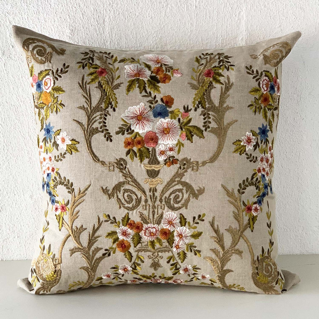 A square-shaped cushion with floral damask embroidery in gold and colorful threads on a natural linen background.