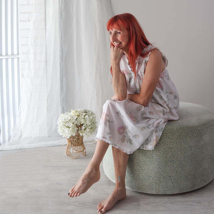 Model seated in natural light wearing Emma nightie, a flowy white cotton gown with soft floral pattern