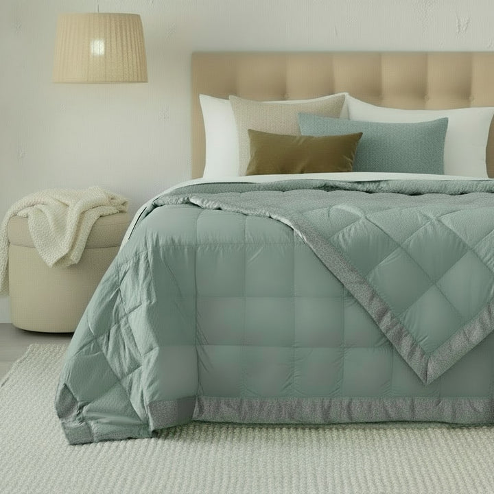 A quilted comforter in a muted sage green color with a gray border, styled on a bed with a beige tufted headboard, white pillows, and accent pillows in olive green and light blue, in a softly lit bedroom setting.