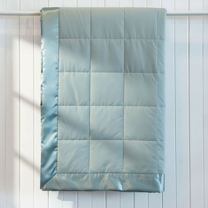 A light blue-green quilted comforter or duvet with a satin border, hanging from a white rod against a white paneled wall.
