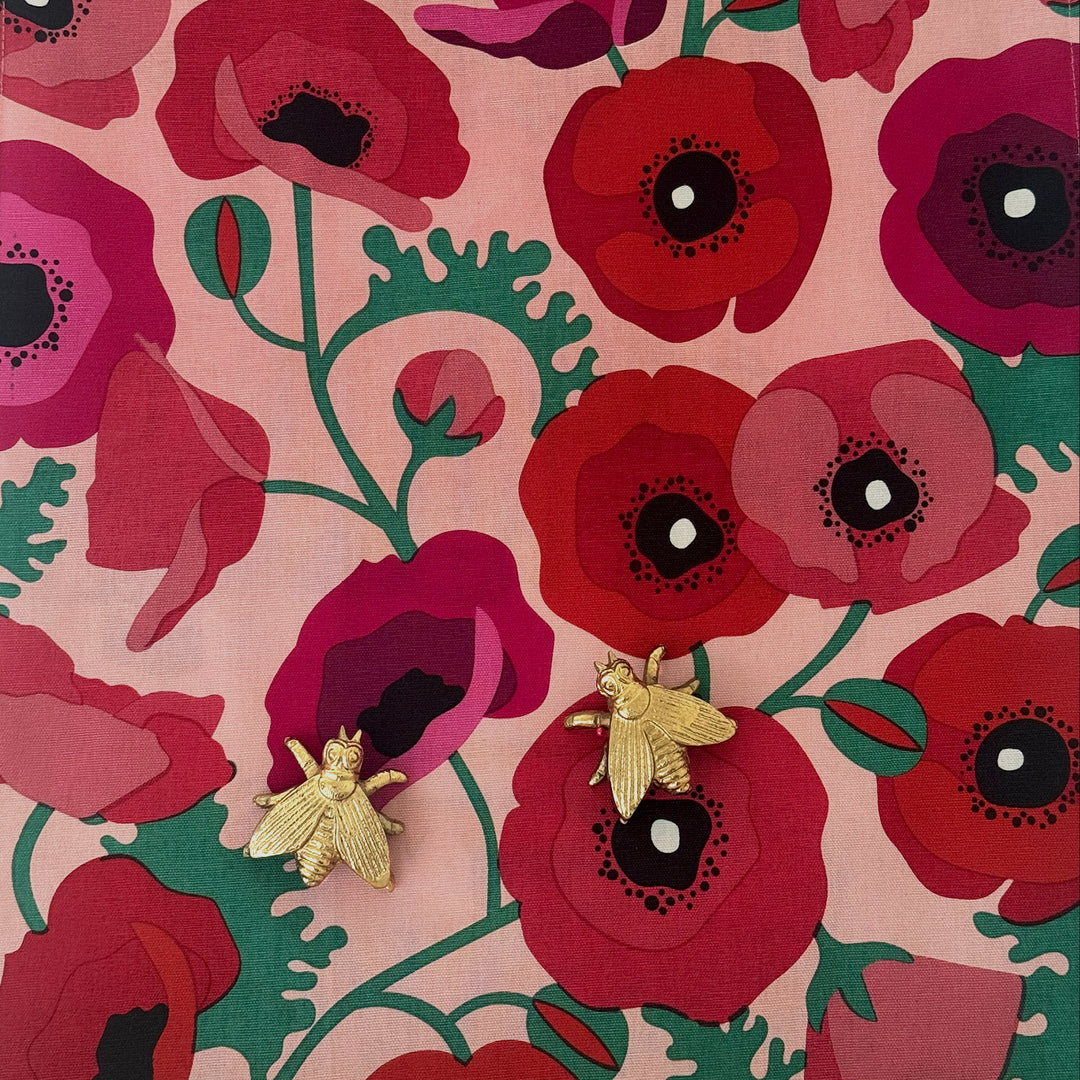 Gold bee earrings on a pink floral background with red poppies and green leaves.