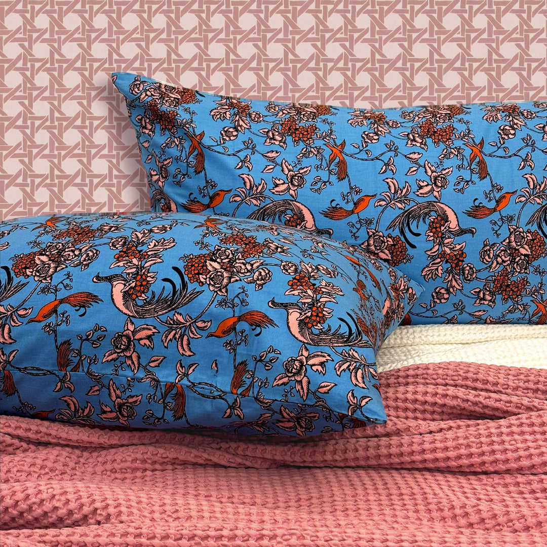 Floral and bird patterned blue pillowcases on a bed with pink bedding and geometric wallpaper.