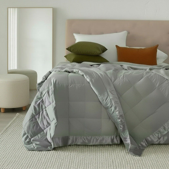 A neatly made bed in a bright bedroom setting, featuring a light gray quilted comforter with a ruffled edge, white pillows, two olive green pillows, and one rust orange accent pillow. A beige upholstered headboard, a large floor mirror, and a cream ottoman are visible.