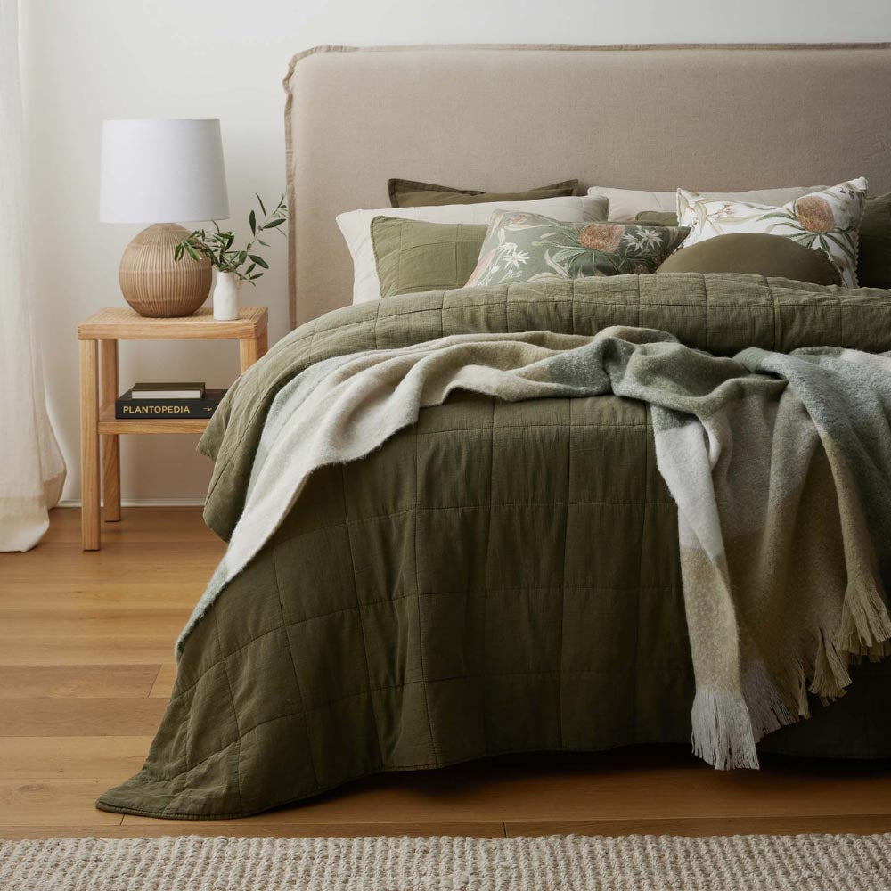 Olive green quilted coverlet styled on a bed with printed cushions and soft wool throw