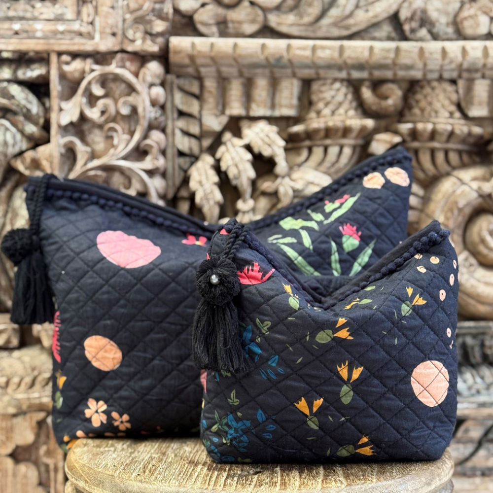 Set of two quilted beauty bags with floral patterns on a black base, styled on a carved wooden background—small make-up bag in front, large toiletry bag behind.