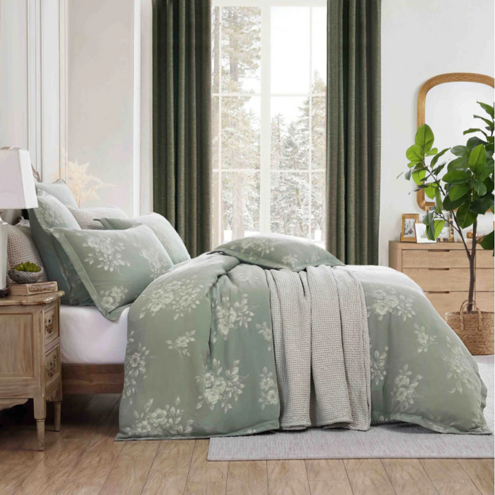 Sage green floral quilt cover on a bed next to wooden drawers, styled in front of a window with green velvet curtains