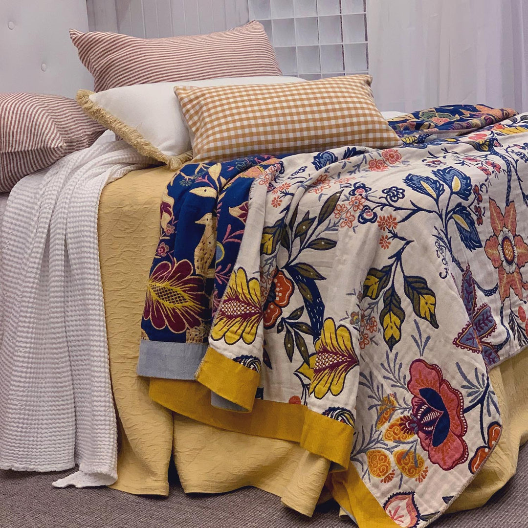Reversible Willow throw styled on bed with floral cream side showing, layered with textured cushions and yellow bedspread.